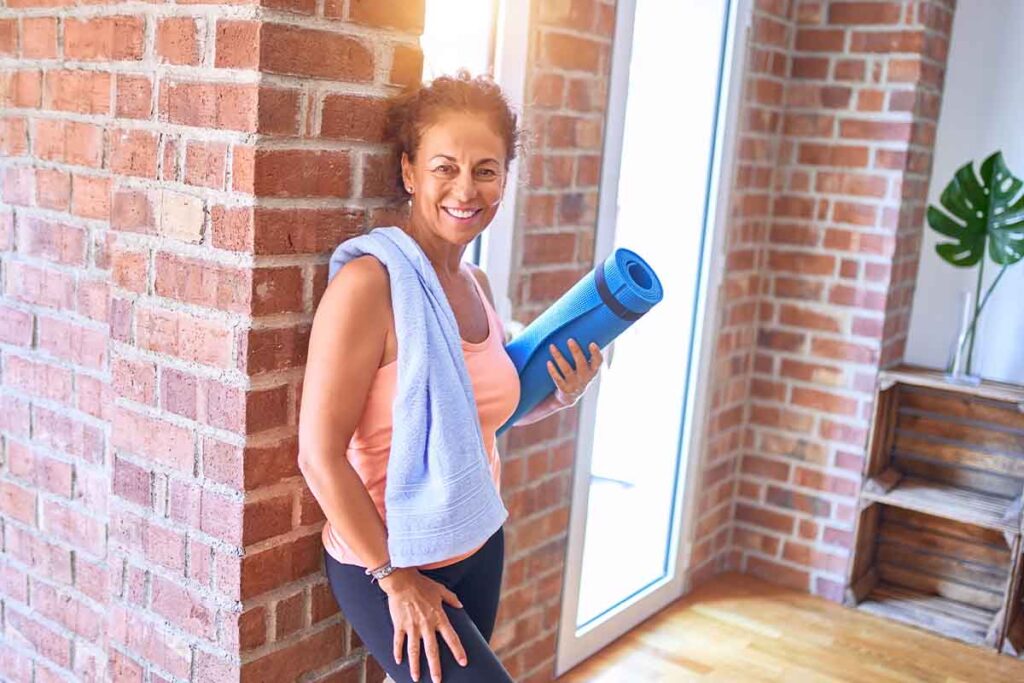 Smiling older woman leaning against wall and holding yoga mat while preparing to do wall pilates for seniors