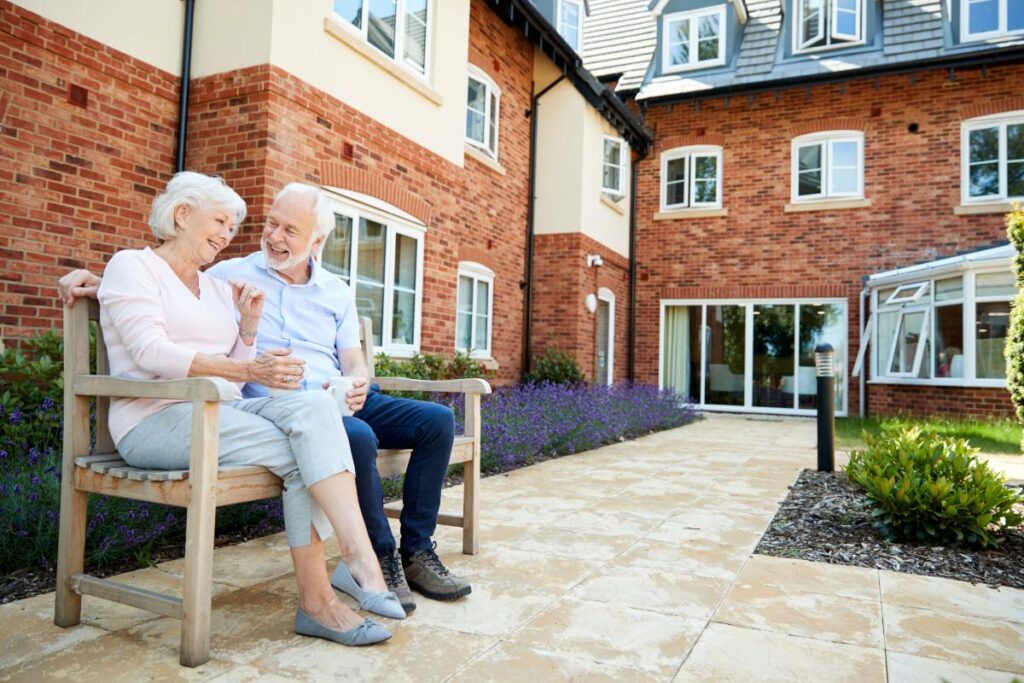 Older couple smiling and laughing while sitting on park bench outside luxury senior apartments in dallas